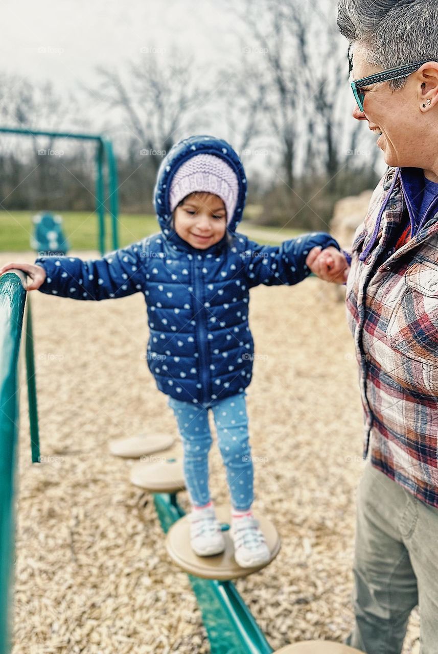 Mother holding hand of toddler girl as she walks across playground climbing bridge, toddler plays at park with mother, having fun with mommy at the park, smartphone portrait photography, picture of happiness, loving moments between parents and child