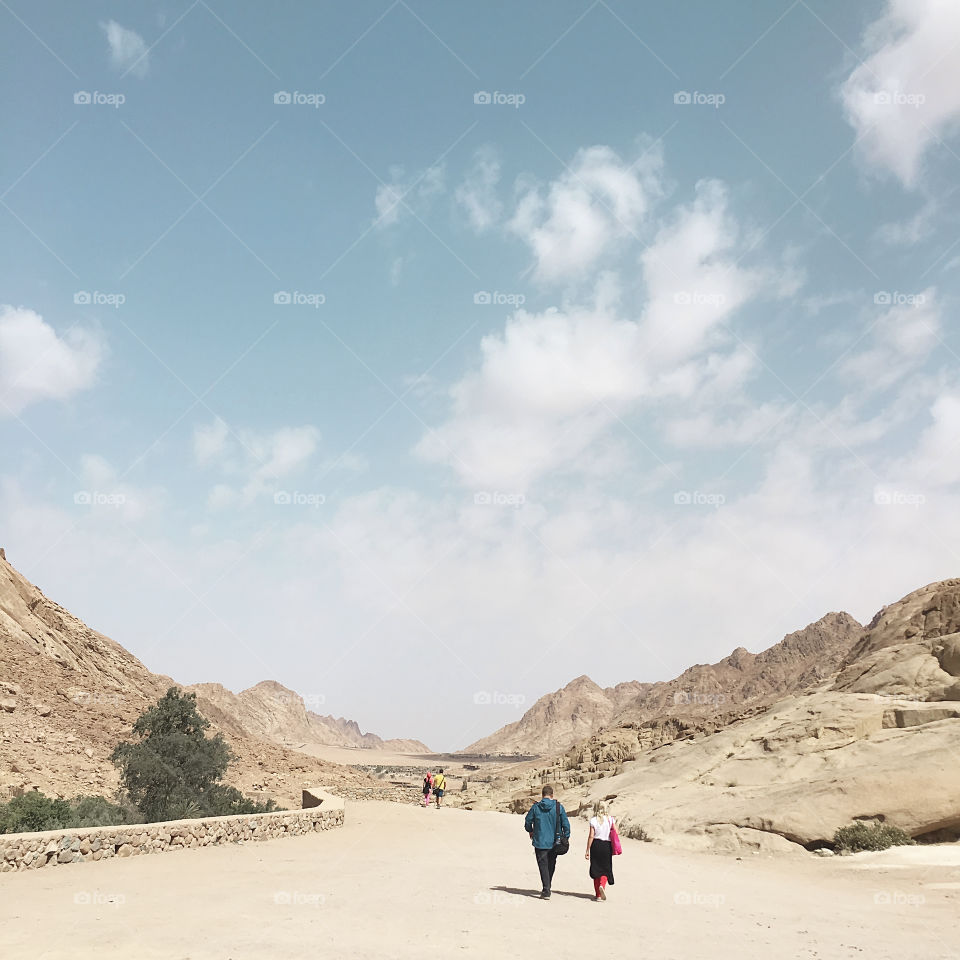 Tiny humans walking by the road surrounded by Rocky Mountains in front of blue cloudy sky 