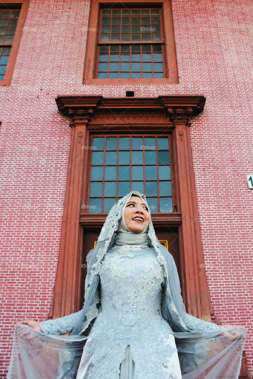 Portrait of young and beautiful Asian muslim woman wearing silver robe with matching hijab and headscarf, holding headscarf and smiling, Red brick wall background. for the wedding.