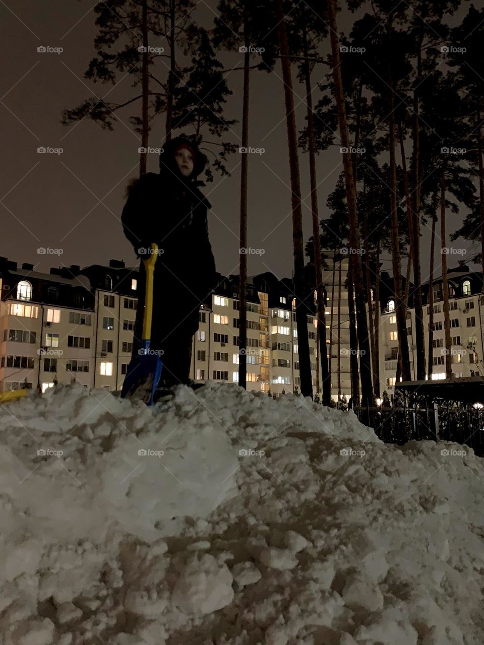 a boy with a shovel stands on a drift of snow against the background of houses with illuminated windows