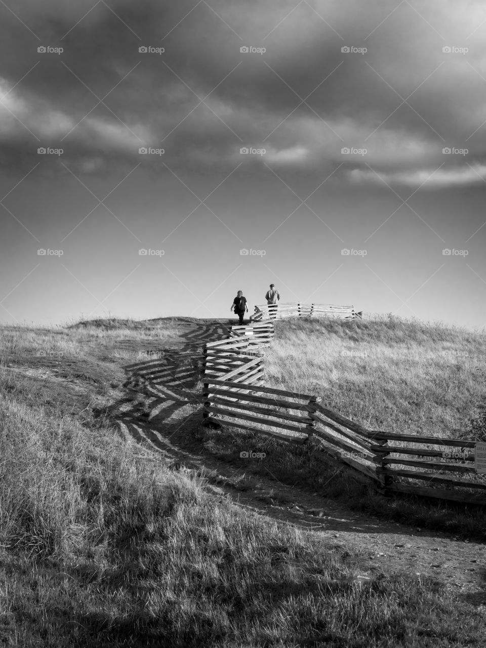 Dark cloud hangs overhead as couple walk up a winding path along a wooden fence