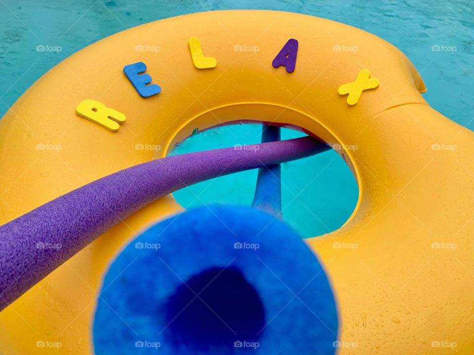 RELAX Word Written On An Eaten Donut Pool Floaty With A Purple And A Blue Floating Pool Noodles.