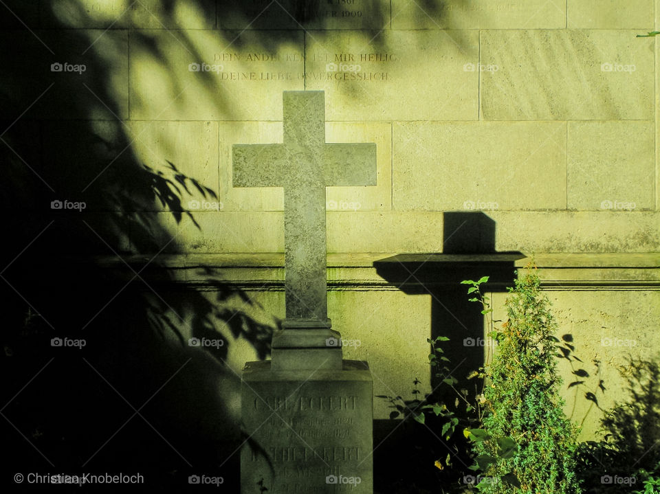 Ein Steinkreuz auf einem Friedhof wirft einen Schatten an eine Wand