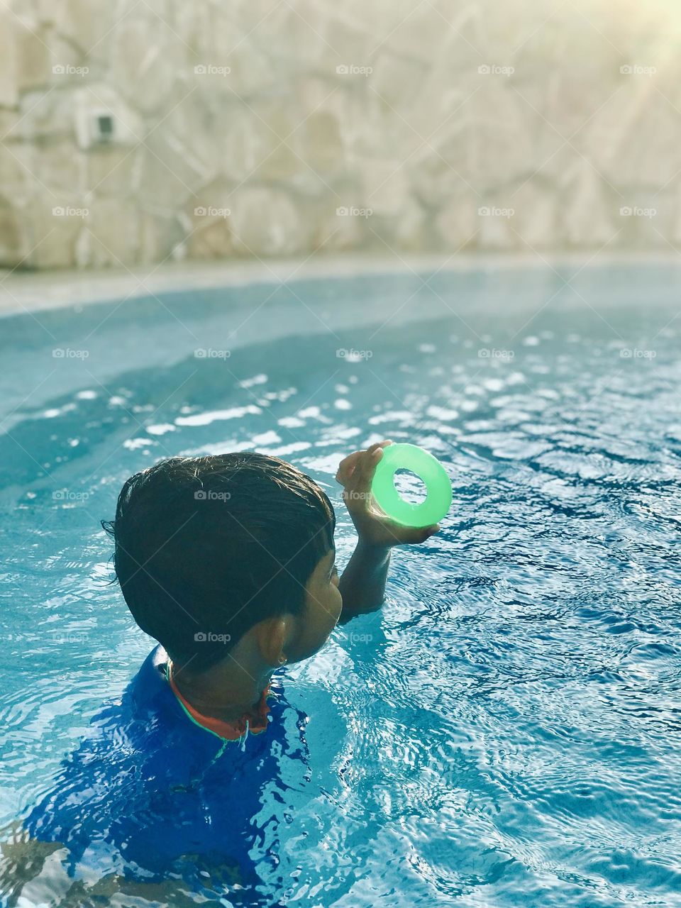 A funny boy looking towards sun through the green circle inside the pool and he’s wearing blue swimsuit ☀️