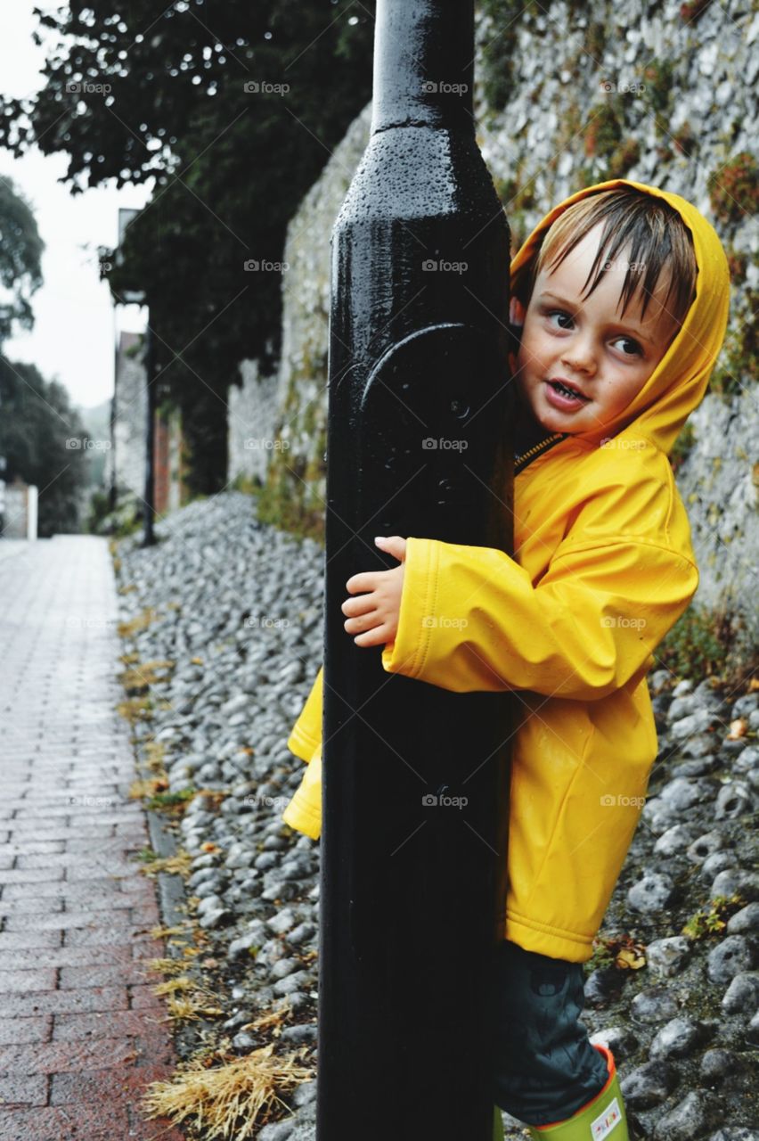 Boy playing in a yellow raincoat in the rain holding on to a street lamppost
