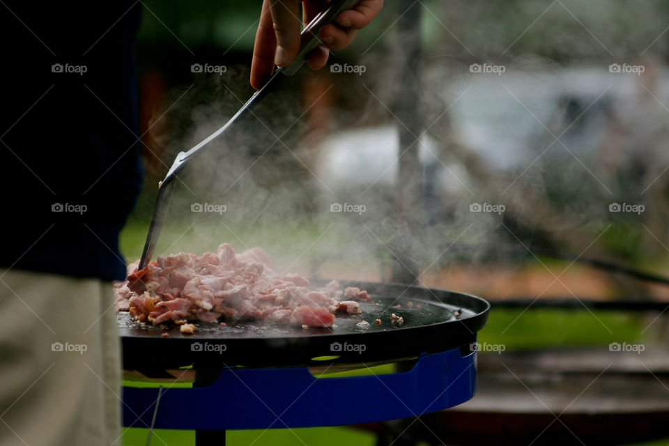 Delicious bacon made outdoors on a gas braai while camping with smoke. Image of man cooking meat outdoors.