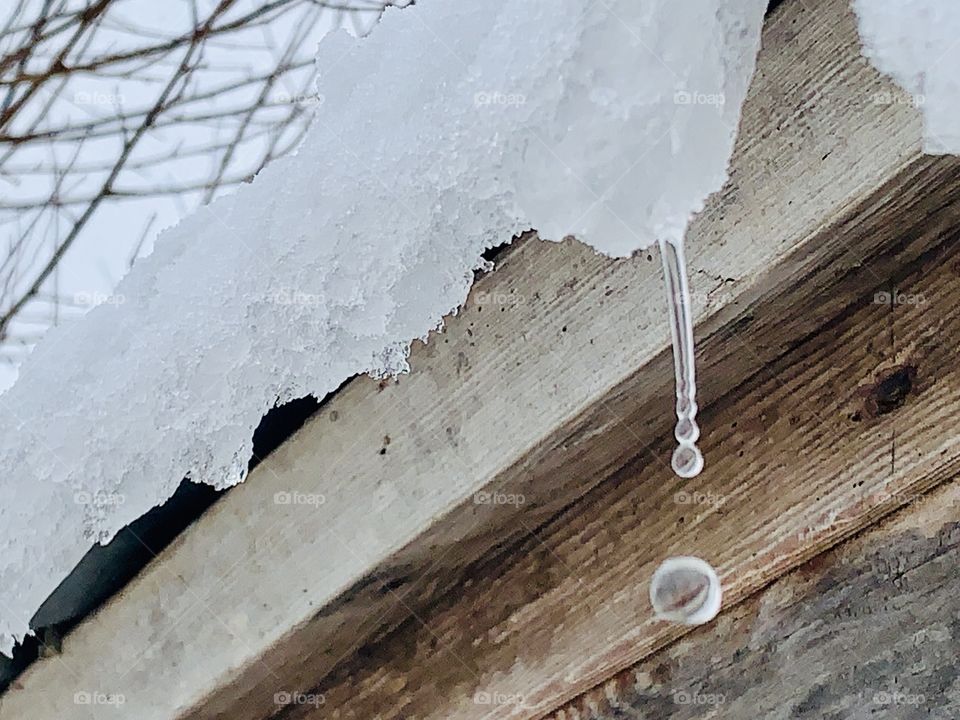 A drop of water falling from melting snow and ice on the roof overhang of a wooden structure 