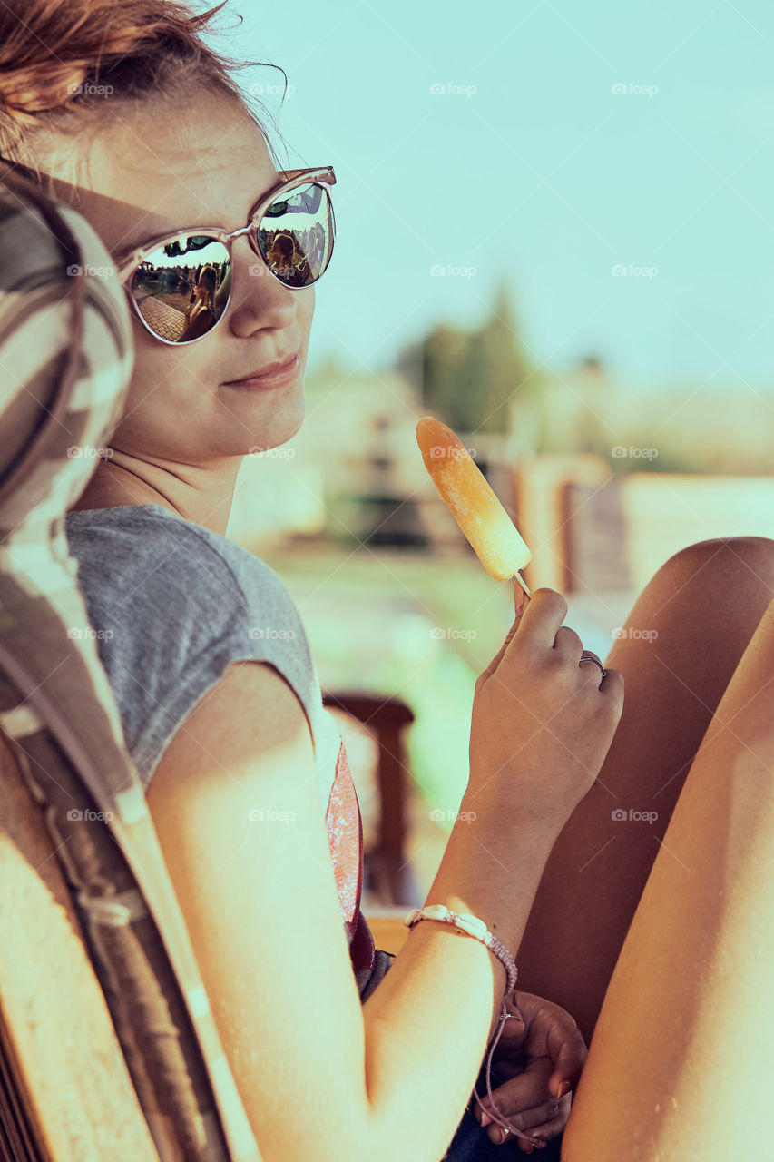 Young woman eating a ice cream, sitting in a chair outdoors on a patio during summer day. Real people, authentic situations