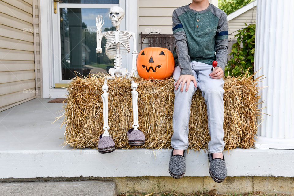 Young boy sitting on a straw bale next to a jack o' lantern trick or treat bucket and sitting decorative skeleton