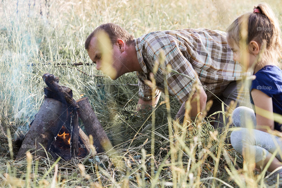 Man starting a campfire, blowing on a fire. Little girl sitting in a grass beside a campfire. Candid people, real moments, authentic situations