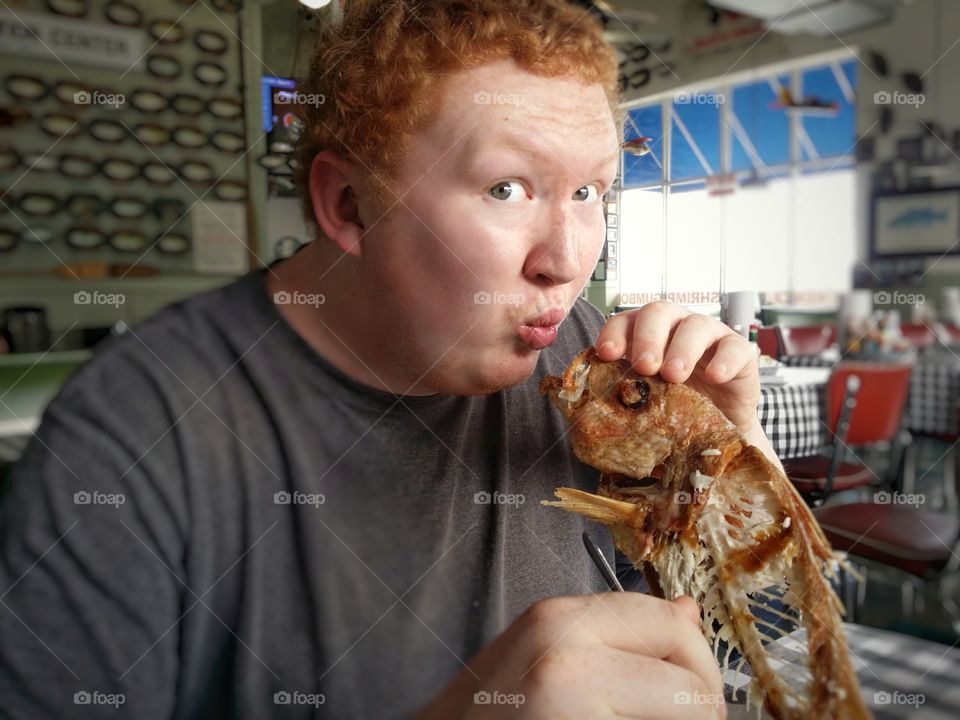 A young redheaded man is sitting in a seafood restaurant pretending to kiss a fried and battered fish that he ate that is now nothing but bones needless to say he enjoyed it...funny :)