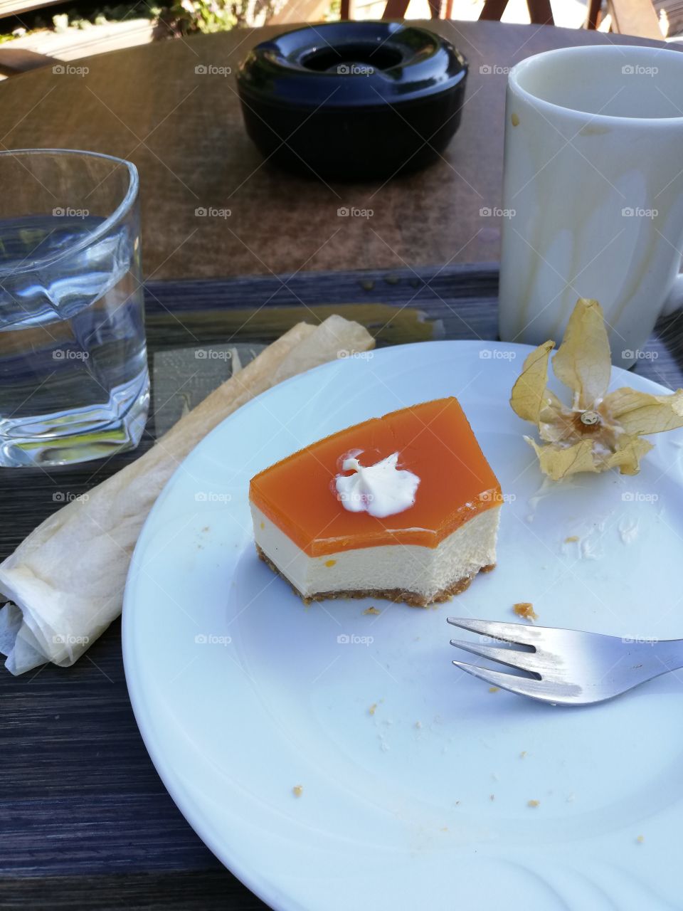 A piece of sea buckthorn berry cake, a fork, crumbs and leaves on a white plate. A glass of water, a messy mug, a wet napkin and spilled milk coffee on a tray. A black ashtray on a table behind.