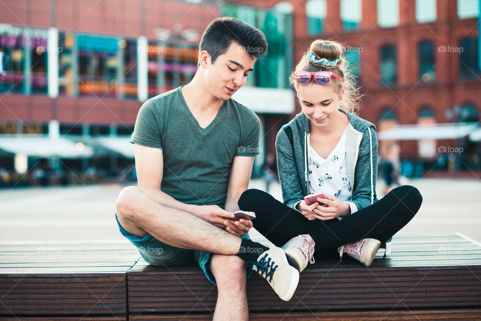 Couple of friends, teenage girl and boy, having fun together, using smartphones, sitting in center of town, spending time together. Real people, authentic situations