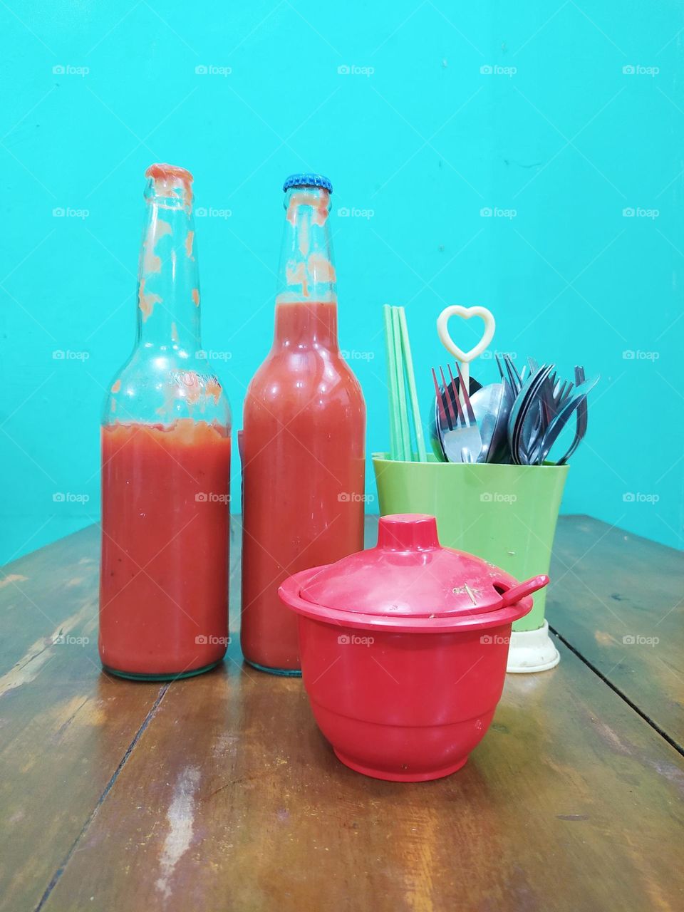 View of the dining table at a local chicken noodle shop, there are bottles of tomato sauce, chili sauce, cutlery, chopsticks on a wooden table with a Tosca wall as a background