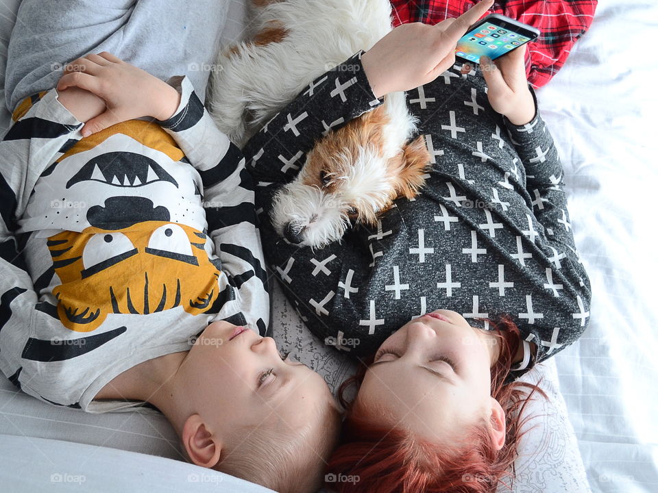 Children using a phone and laying in the bed together with their dog