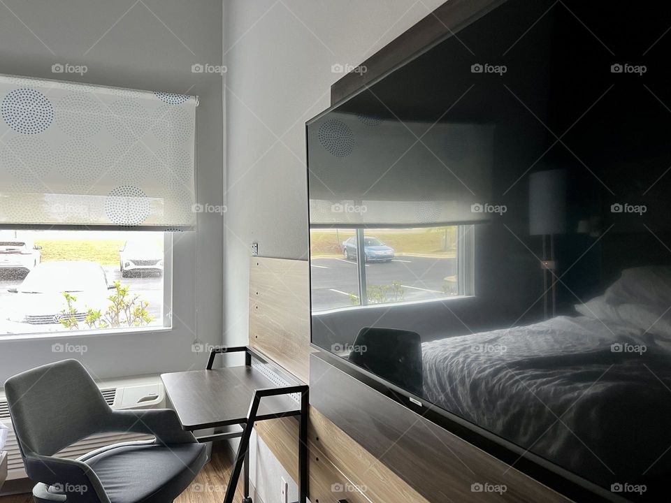 American motel room morning. The ground floor window illuminates a spare desk and chair, while the white linens of a rumpled bed reflect in a large screen wall mounted television.