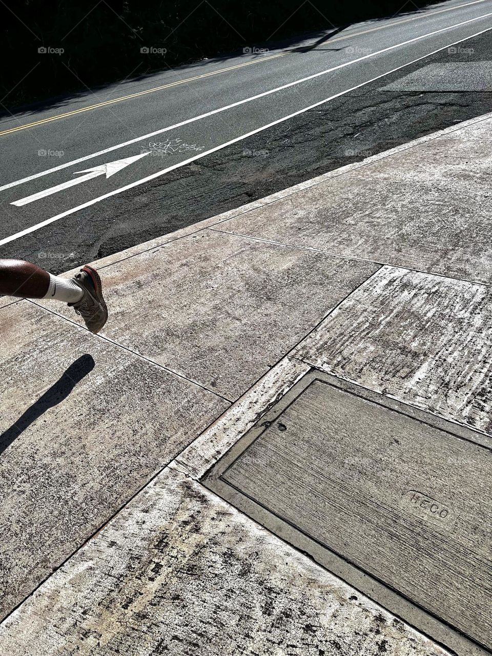 Just a glimpse of a lower leg and foot of a man running downhill on a sidewalk near a bike lane and road