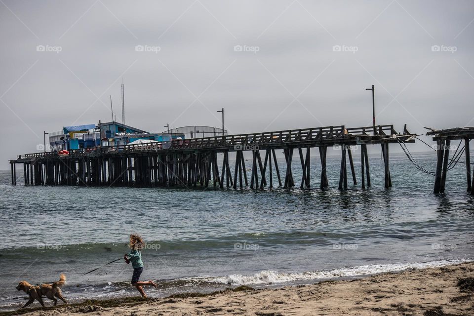 Young girl walking her dog on the beach with the ocean waves on a beautiful day in Capitola California with the wharf in the background