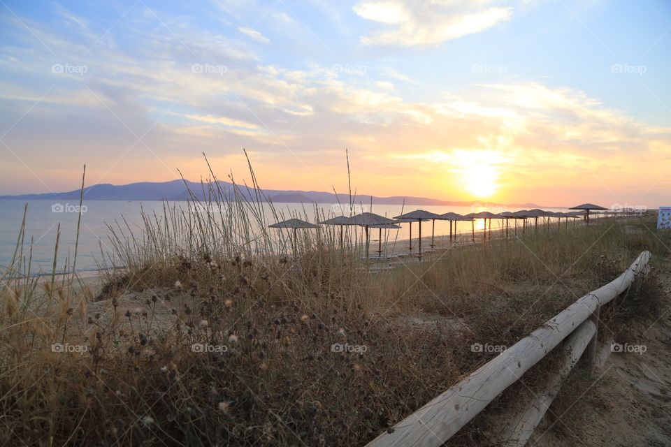 When the sun is about to go down behind the mountains in front of the sandy beach with a row of parasols standing in the foreground seen from the road where I’m taking the photo from my perspective 