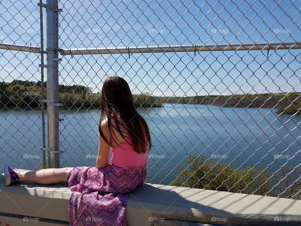 Rear view of a girl sitting near the fence looking at lake