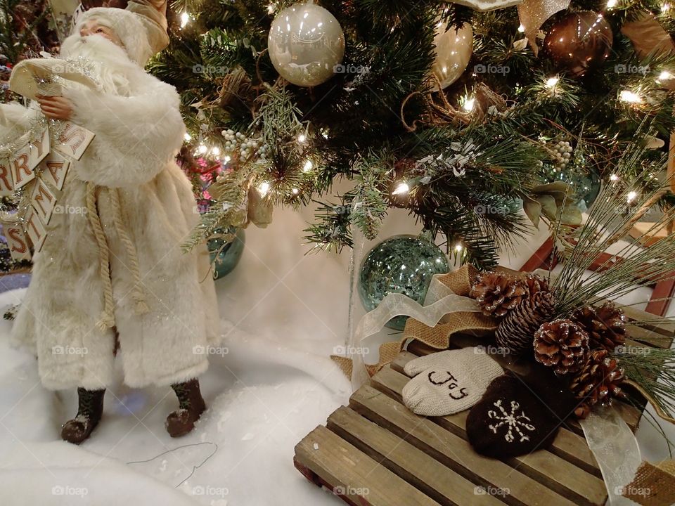 A white clad traditional Santa Claus holding a “Merry Christmas” sign at the base of a lighted Christmas tree. 