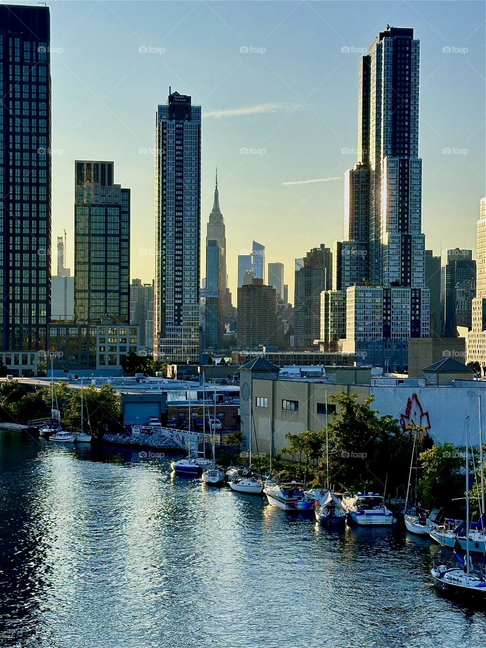 This is the LIC shore at “Newtown Creek” up close with its many boats seen from the “Pulaski Bridge” that connects “Greenpoint”, Brooklyn to LIC, Queens. In the distance we see “Manhattan” and the “Empire State Building”. 2024. Hypnotic Productions