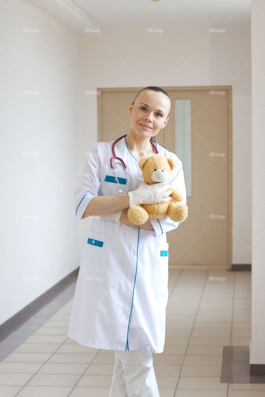 Young pediatrician in a professional uniform with plush teddy bear stays in the corridor of a hospital.