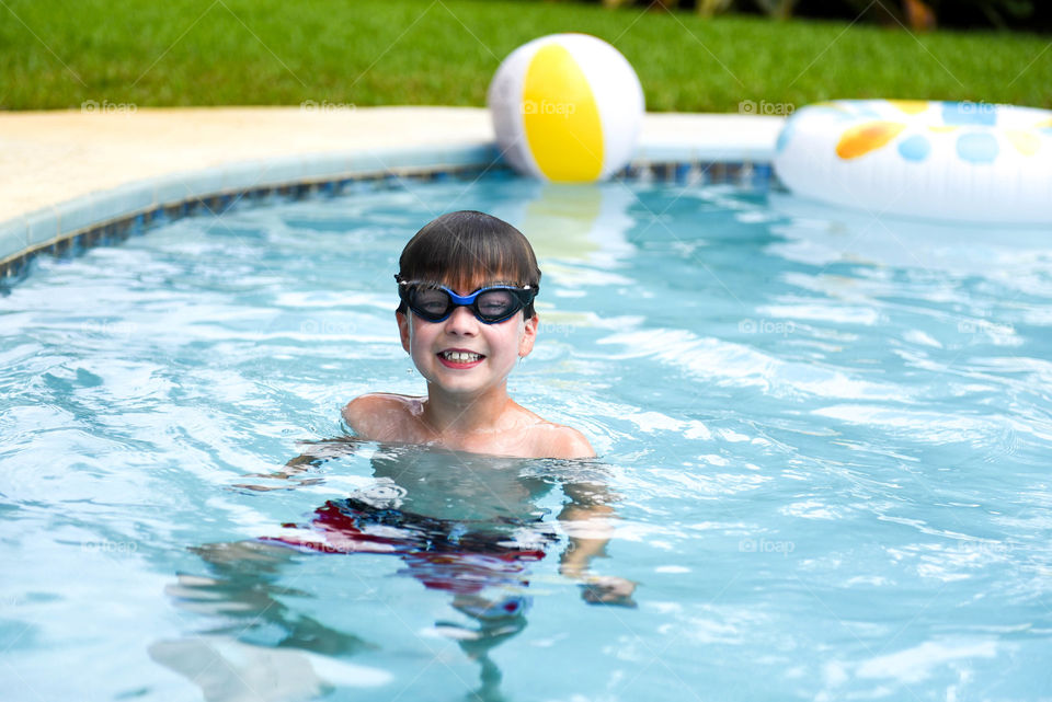 Young boy with goggles on smiling and swimming in a pool outdoors