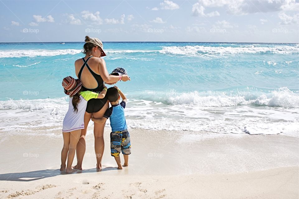 Mom and her 2 children, all in swimsuits and hats, stand on a beach shoreline facing the water. As 1 child hugs her, the other holds tight to her leg.