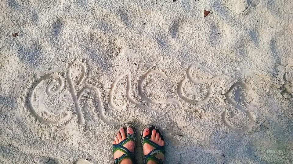 Feet wearing Chacos and covered in white sand at the beach with the word "Chacos" written in the sand above them.