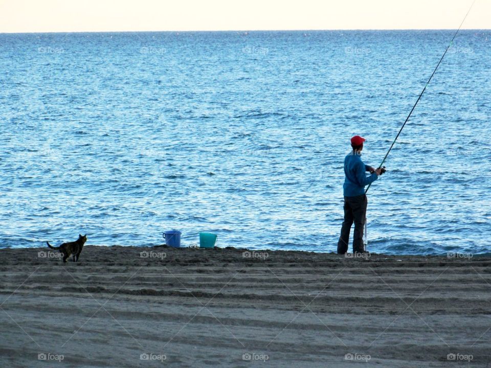 Fisherman on the beach and cat. Fisherman fishing on spinning on the beach of sea. Nearly are two buckets. A cat sneaks up as a hunter from behind.