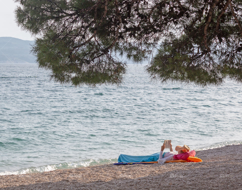 Summer vacation alone, relax. The girl lies on a deserted pebble beach at the edge of a calm sea under the branches of a pine