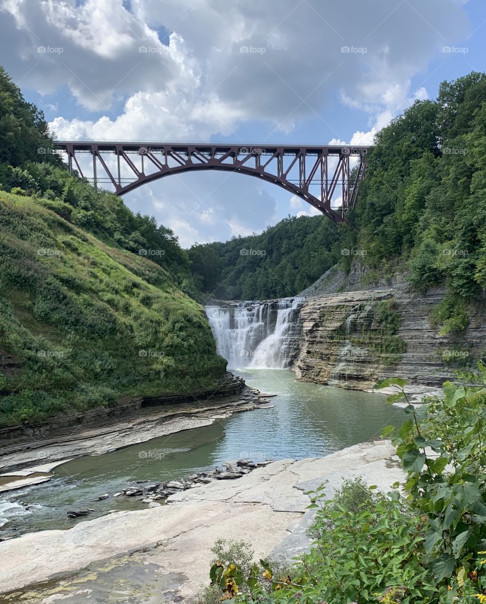Clear photo of a bridge over a river and waterfall. Has beautiful plants and sky with nice complimenting colors and scenery. 