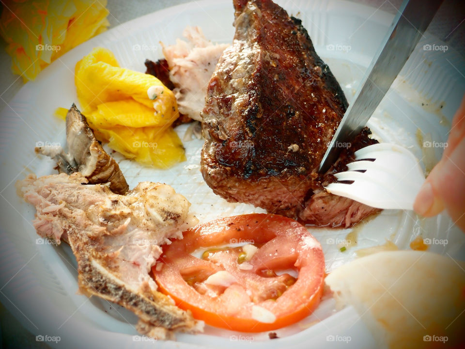 Taking a slice from a steak . A fork and and a knife in a plate, cutting off a piece from a steak. In plate bones and a tomato.