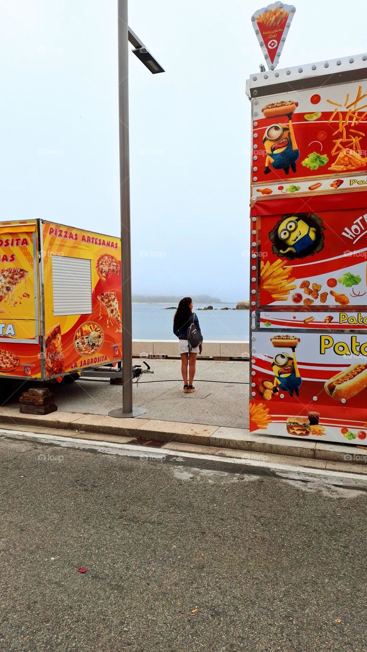 A girl between food trucks, watching the sea, the morning after the village carnival.  Palmeira, Spain.