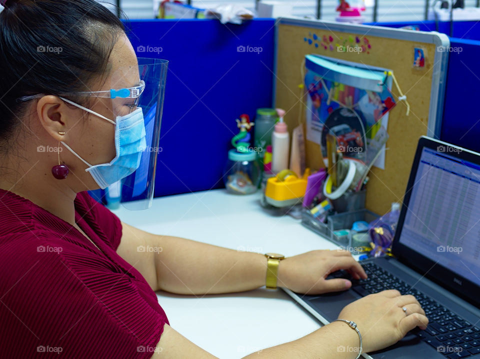 Young woman wearing surgical mask and face shield while working alone inside an office.