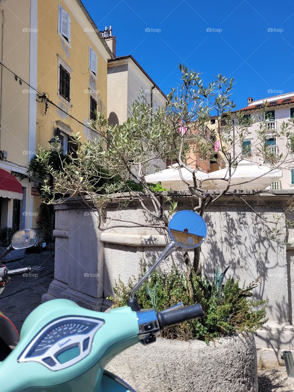 Turquoise Vespa parked in city under intense blue sky also appearing in rearview mirror