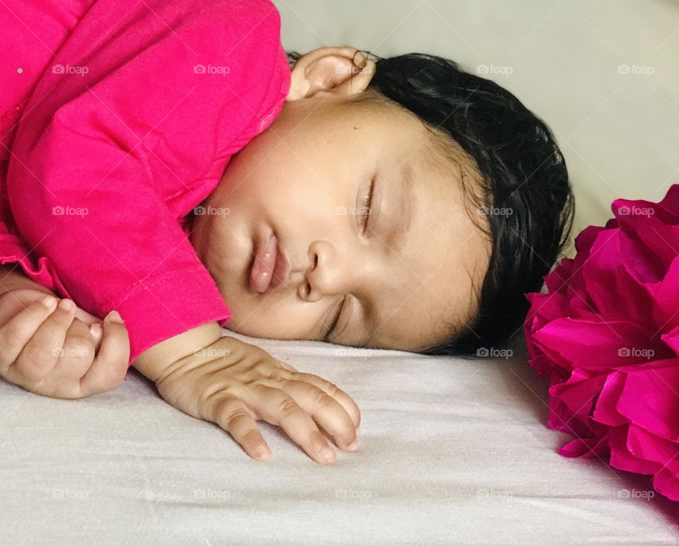 Baby girl sleeping on white blanket and wearing pink dress beside her a pink paper flower 