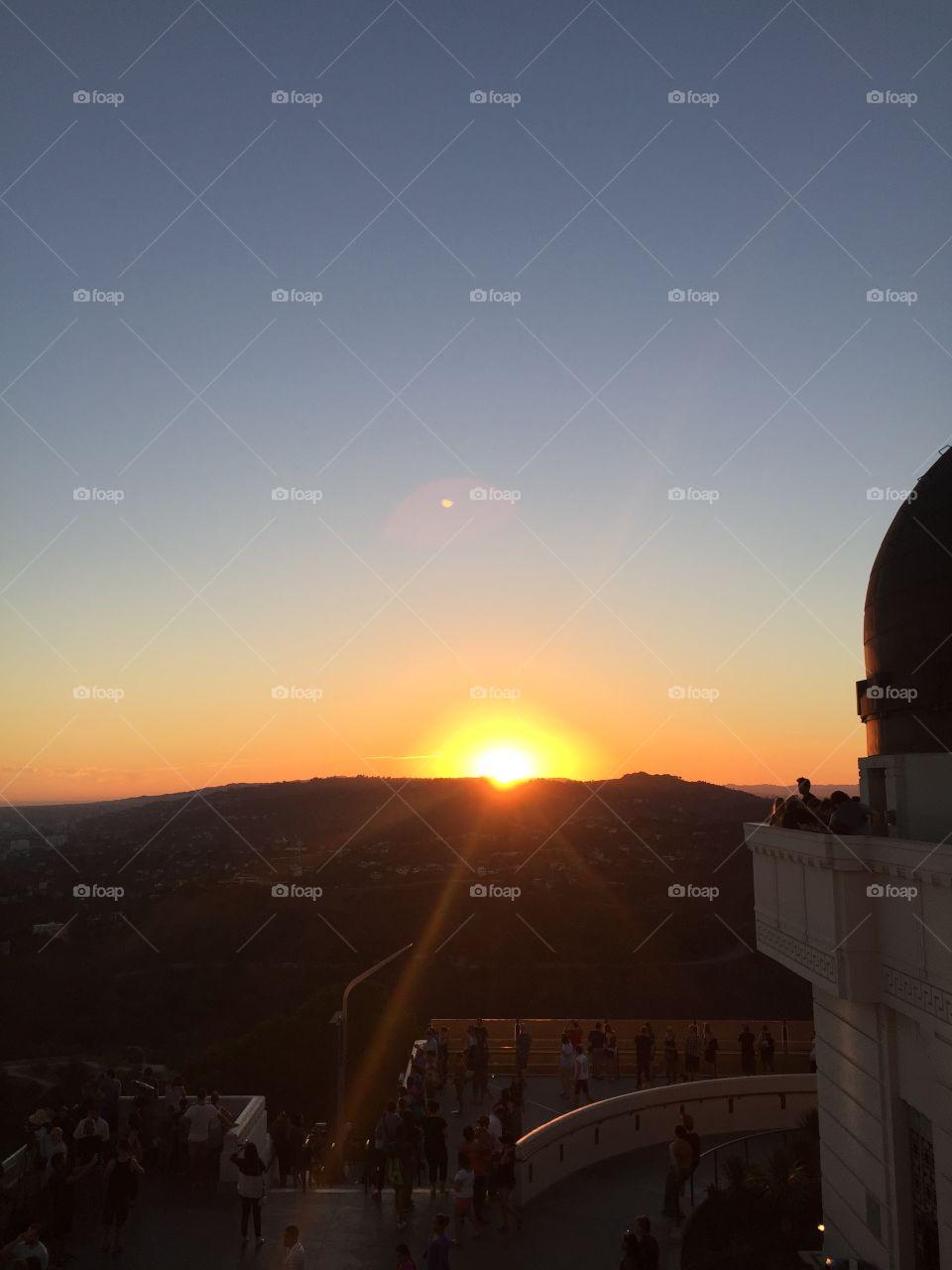 A view of the sun going down behind a mountain against a blue cloudless sky while against the Griffith Observatory.
