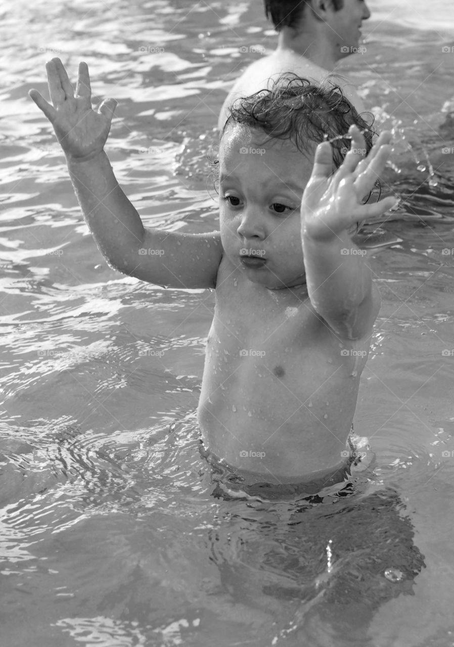 A toddler boy plays and splashes in an outdoor swimming pool on a sunny summer day. 