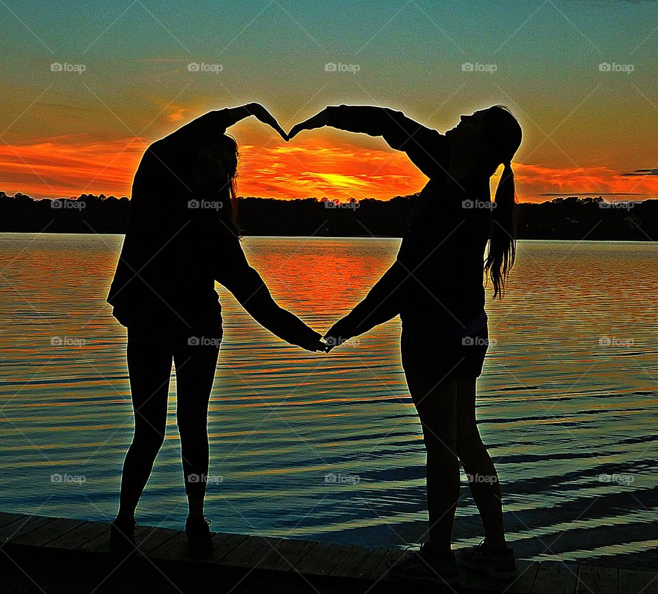 Silhouettes and shadows - Two friends use their arms and body to make a heart with enclosed  fantastic sunset