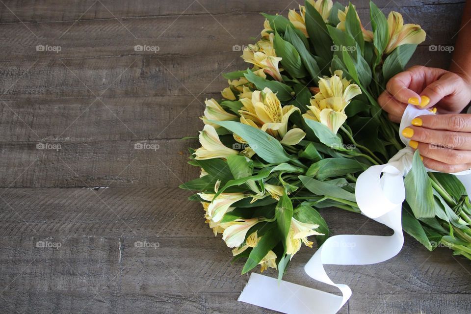 Woman tying ribbon around Bouquet of yellow flowers with yellow nail polish on nails wooden background  