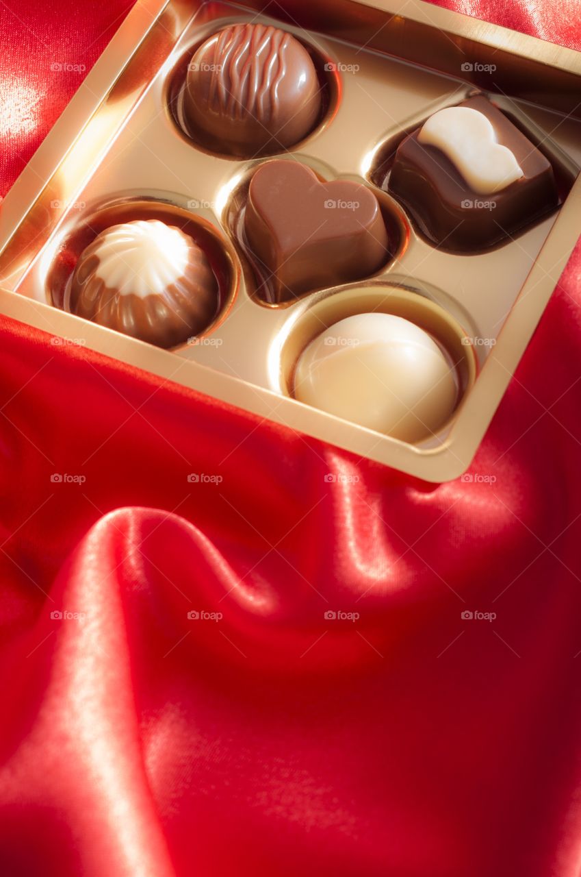 Golden box of differently shaped chocolates on silk background surrounded by rose petals for Valentine's day 