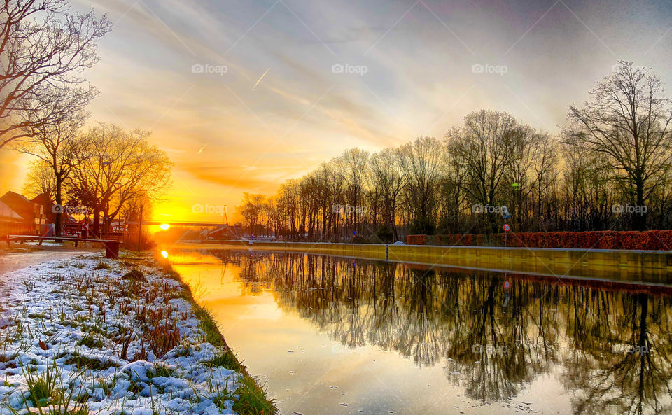 Colorful sunrise or sunset over a winter snowy landscape, reflected in the water of the canal or river