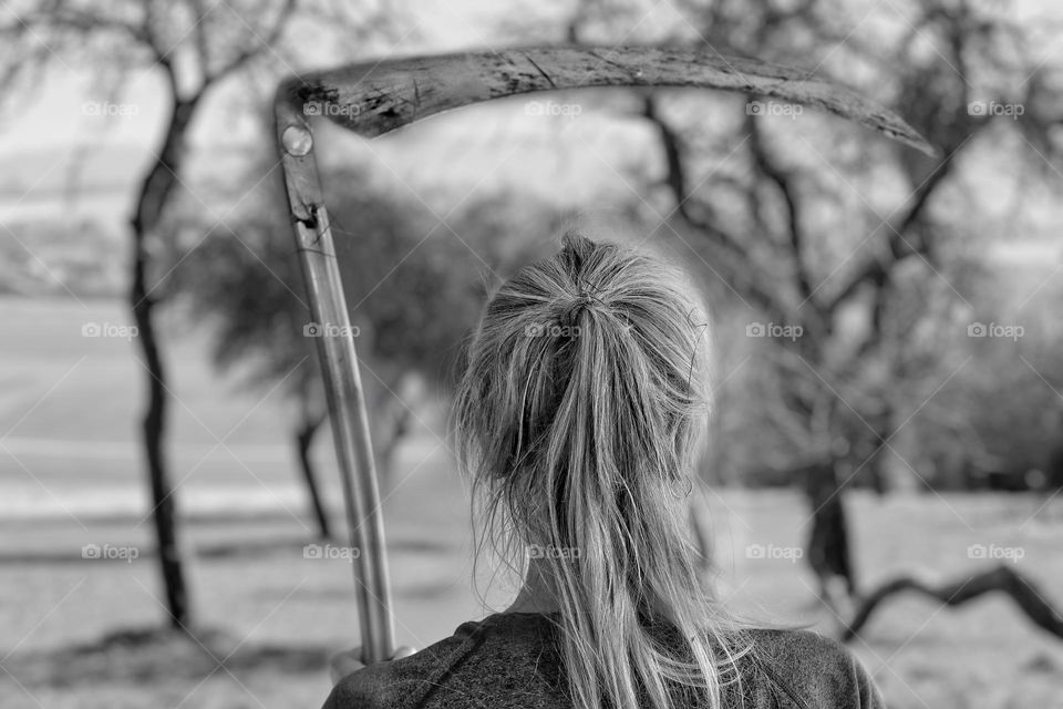 Black and white photo of rear view of a woman holding a scythe in a landscape with trees