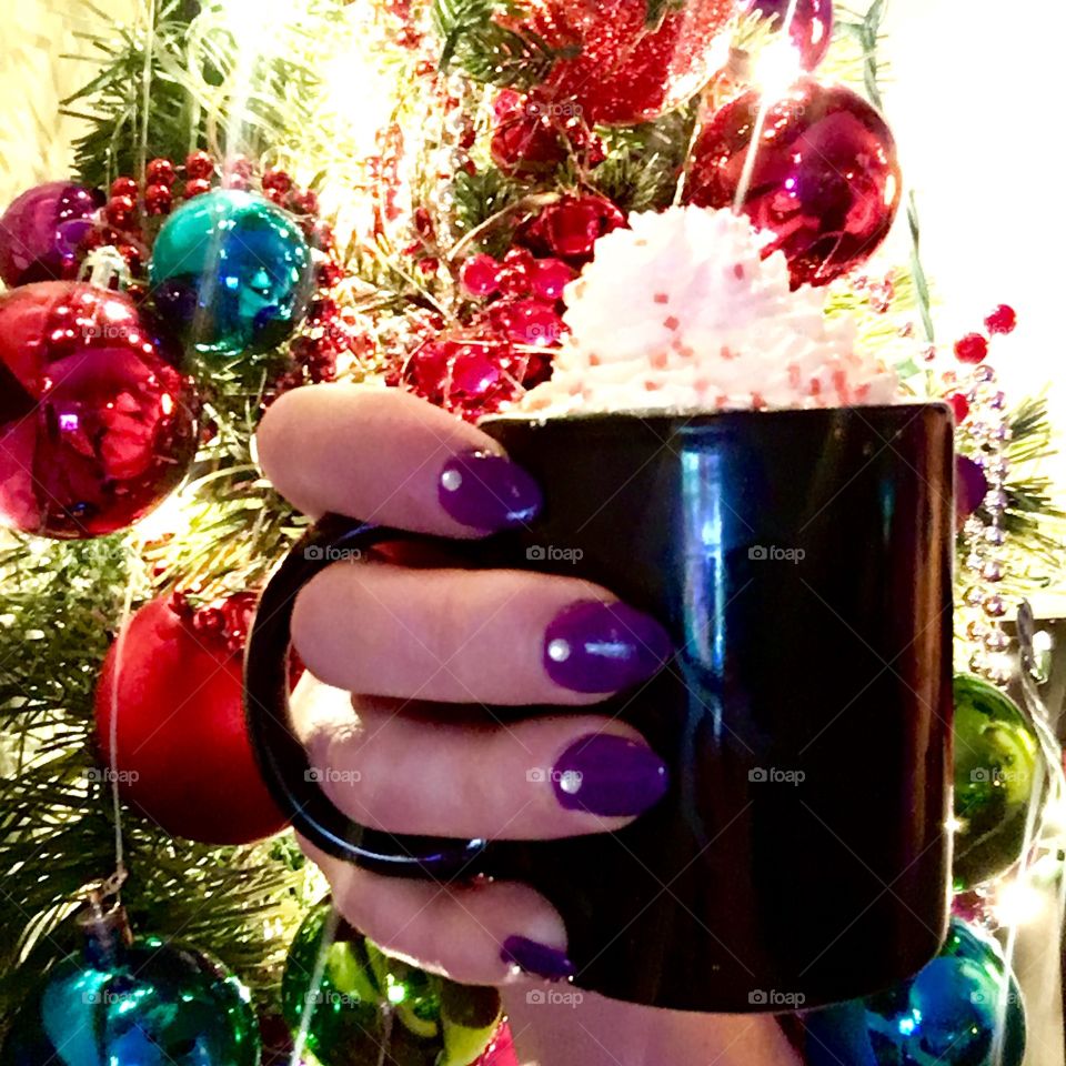 A manicured hand holding a cozy mug topped with whip cream with a festive, textured Christmas background 