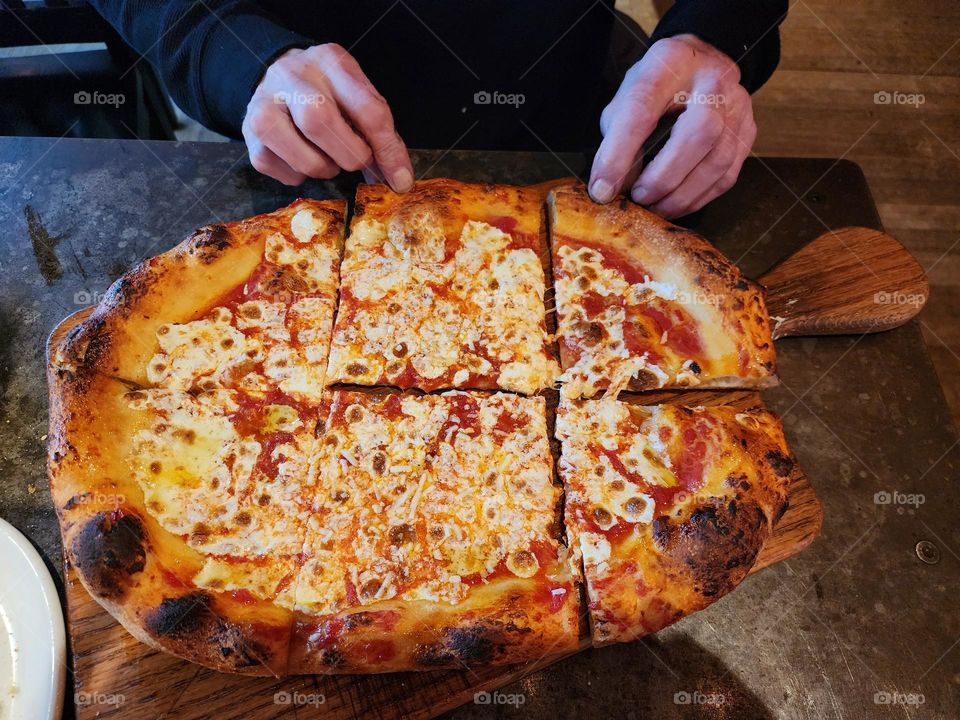 A large cheese pizza is seen from above, looking down at the table and pizza. A man's hands, reach to get a slice.