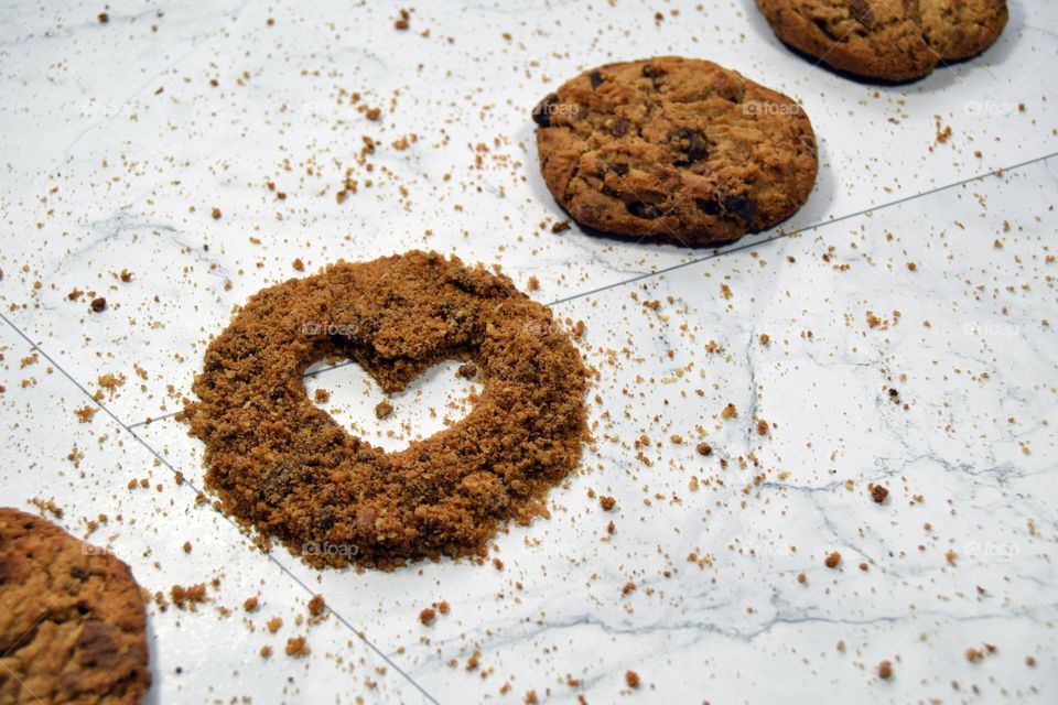 A diagonal line of golden brown chocolate chip cookies featuring a heart hollowed out of a small circular pile of cookie crumbs on a white marble background.