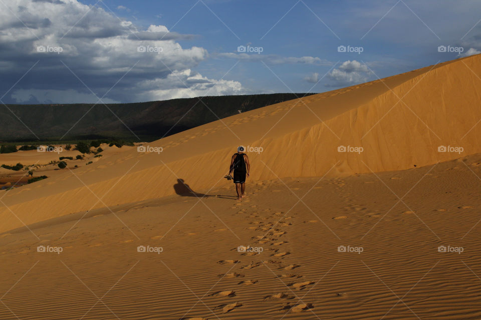 boy walking trhough a big bank of sand, leaving his steps behind him, under a blue beautiful sky and a part of a mountain in the background