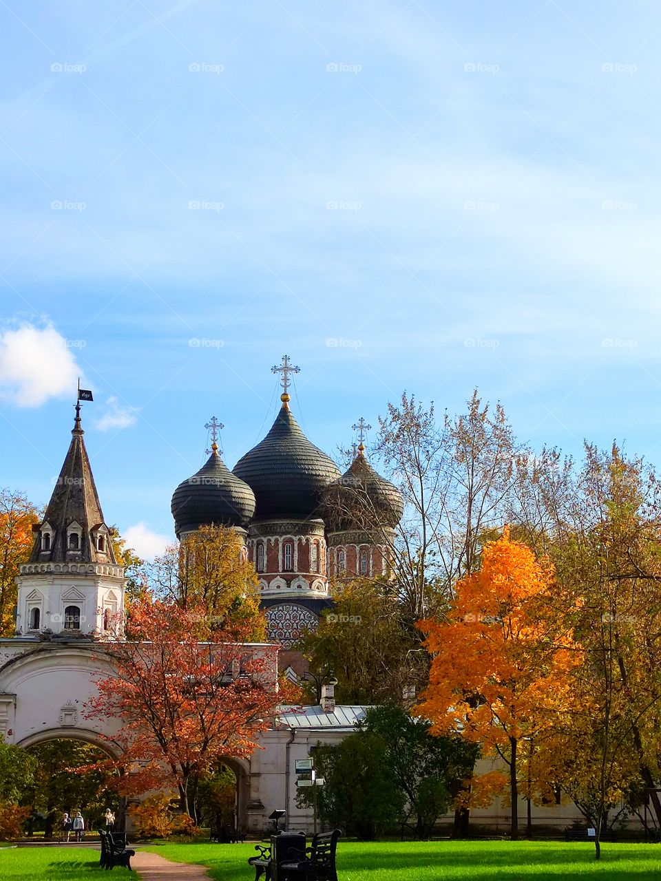 Green lawn with benches.  Multicolored autumn trees.  Ancient fortress wall with a white tower. Domes of an ancient church. Blue sky